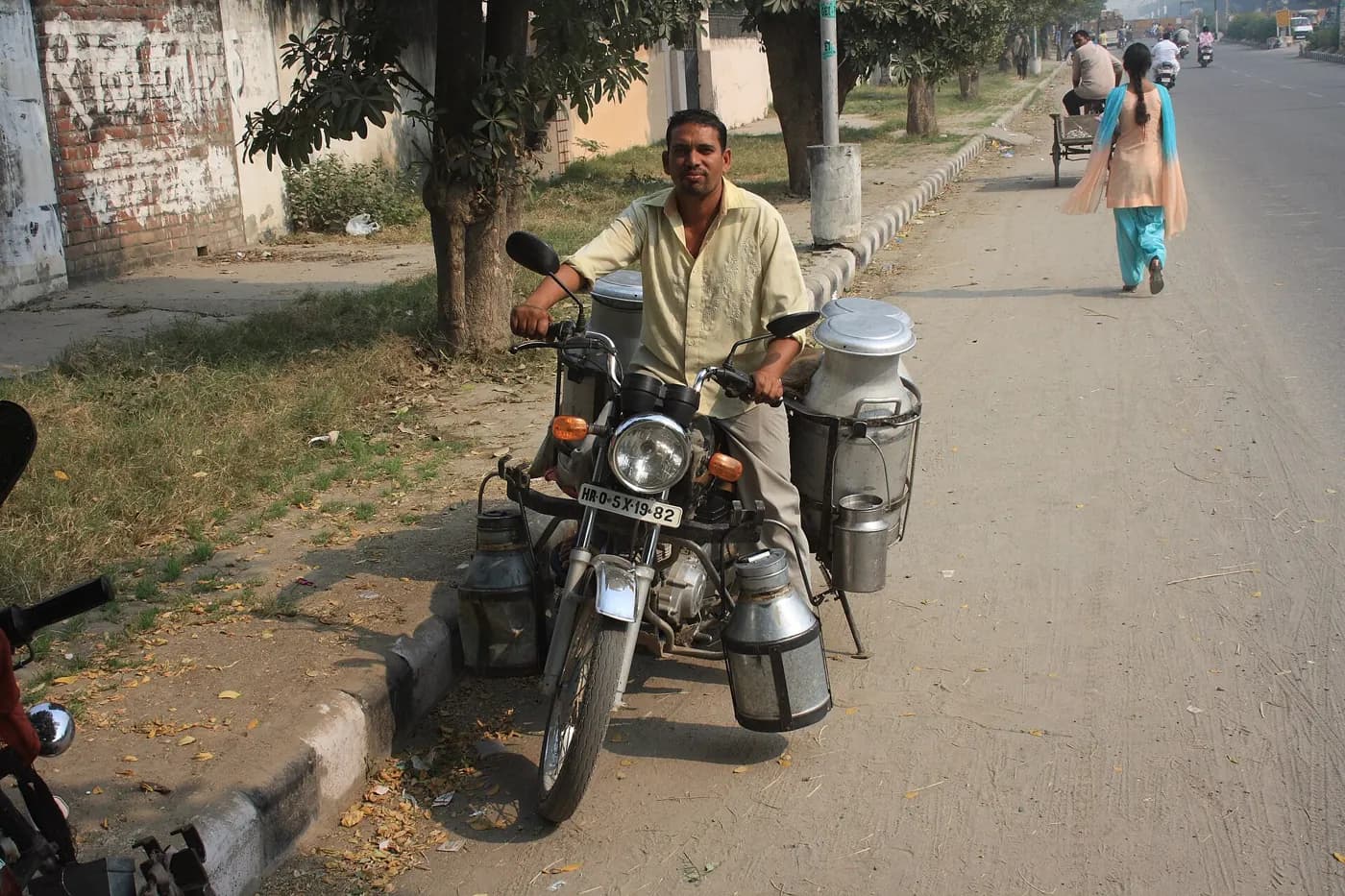 A milkman on a motorcycle hung with steel milk churns, stopped at the side of a road