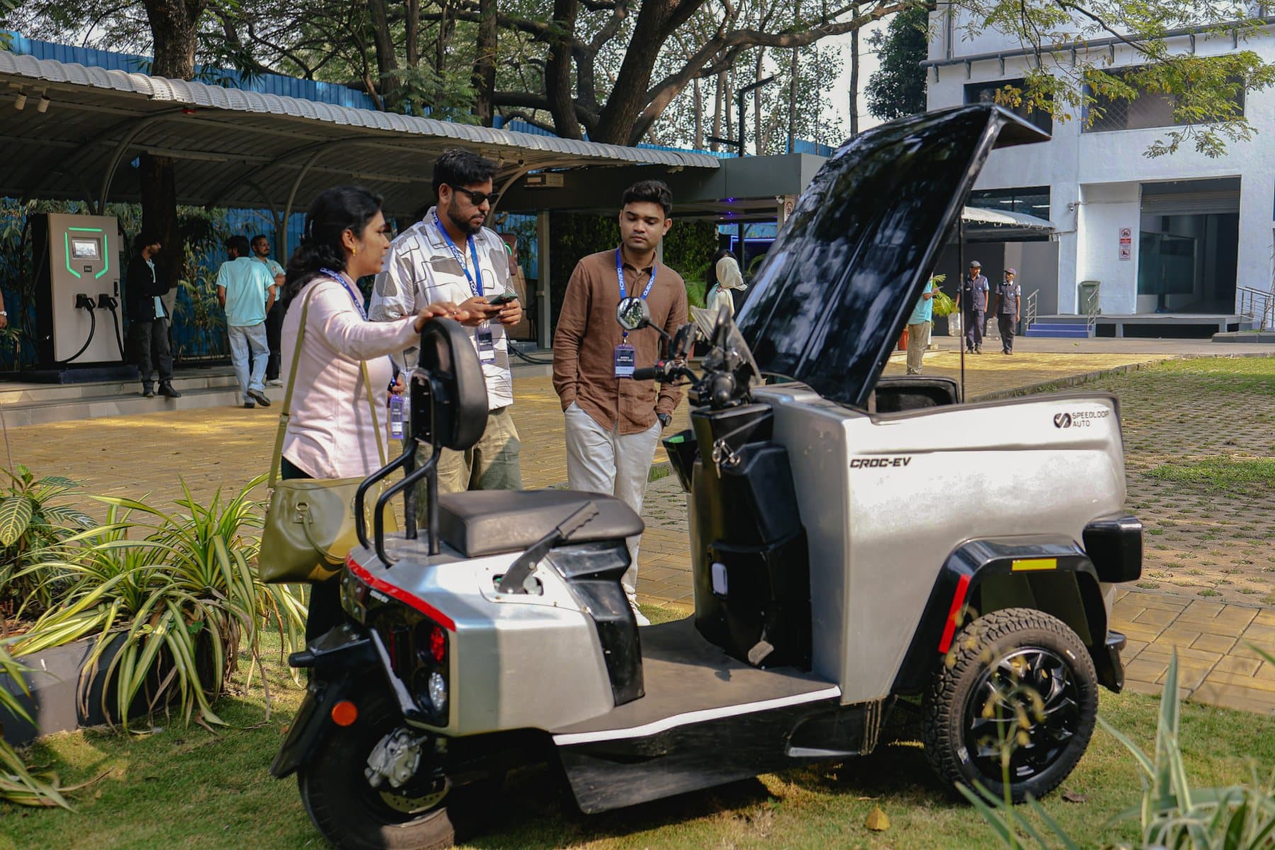Three visitors with lanyards look over a silver CROC EV with its cargo box lid raised, parked on grass at an outdoor expo.