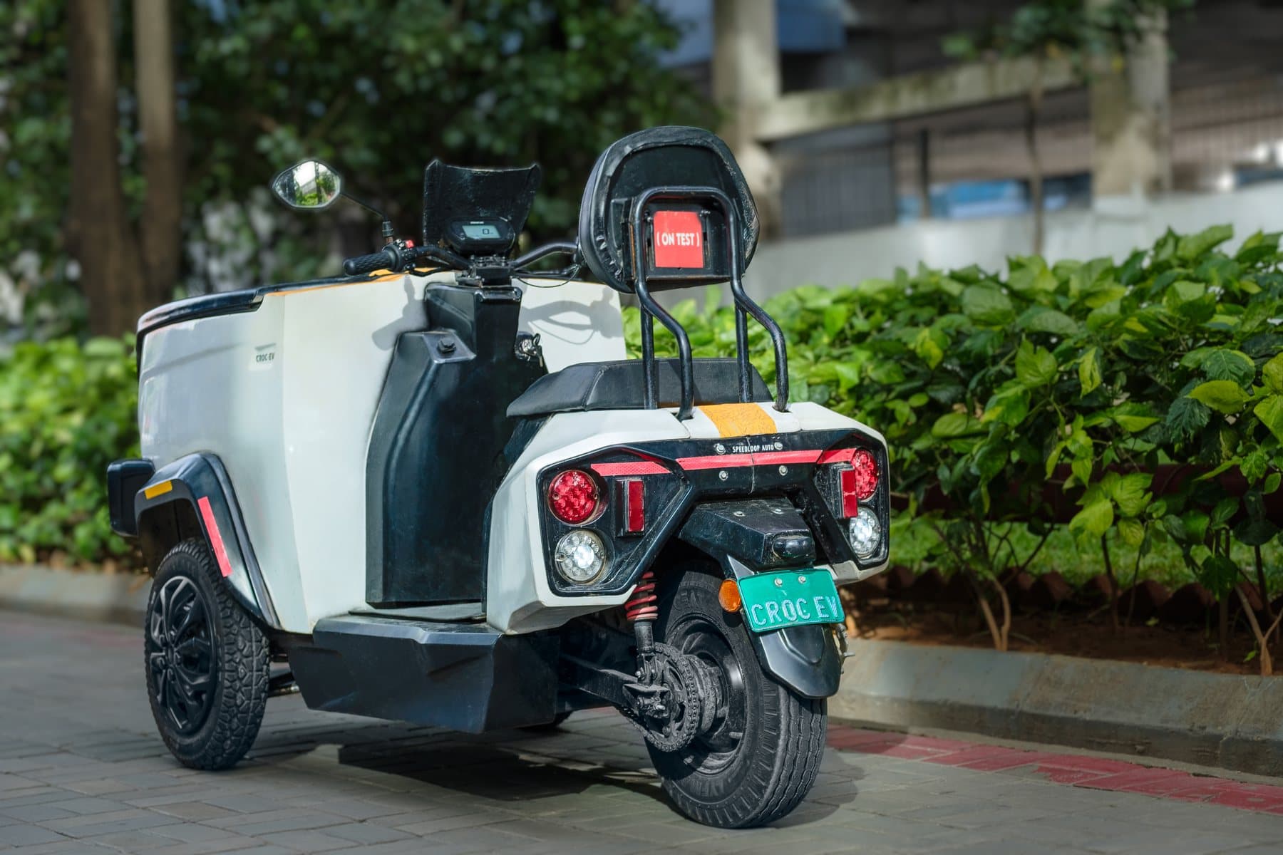 A white CROC EV parked beside a hedge, seen from behind, showing its single rear wheel, tail lamps, rear suspension and chain drive.